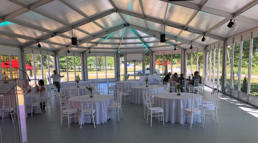 Glass-walled wedding structure tent interior with white chiavari chairs, round tables, and colorful uplighting