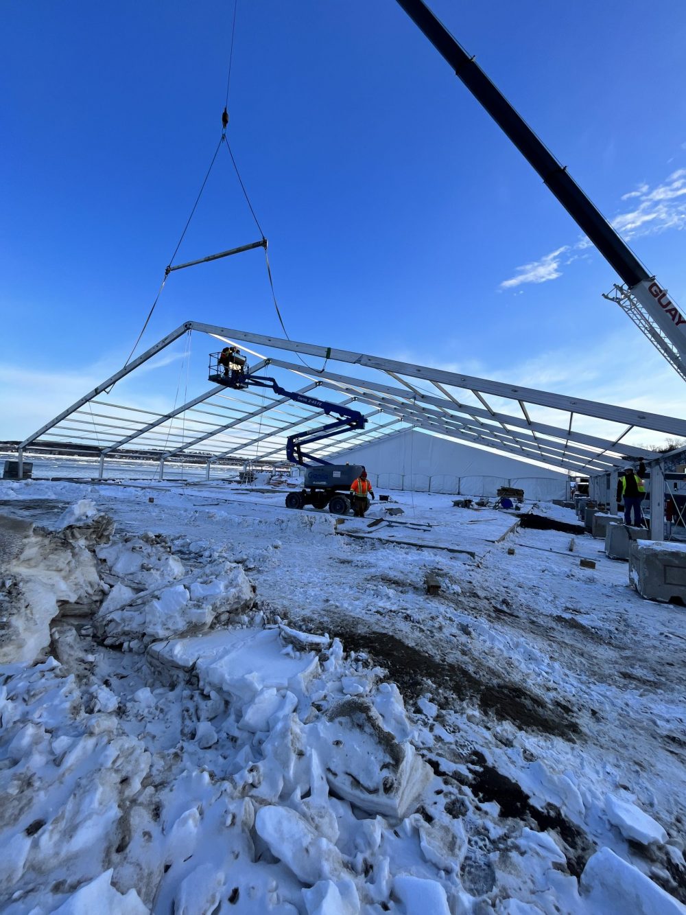 Crane lifting aluminum building restoration tent frame into position during winter installation with snow on the ground