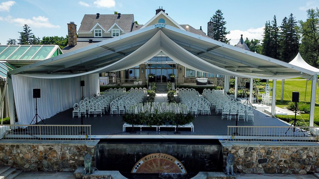 Aerial view of outdoor wedding ceremony with draped structure tent, white chairs, and floral arrangements at luxury estate