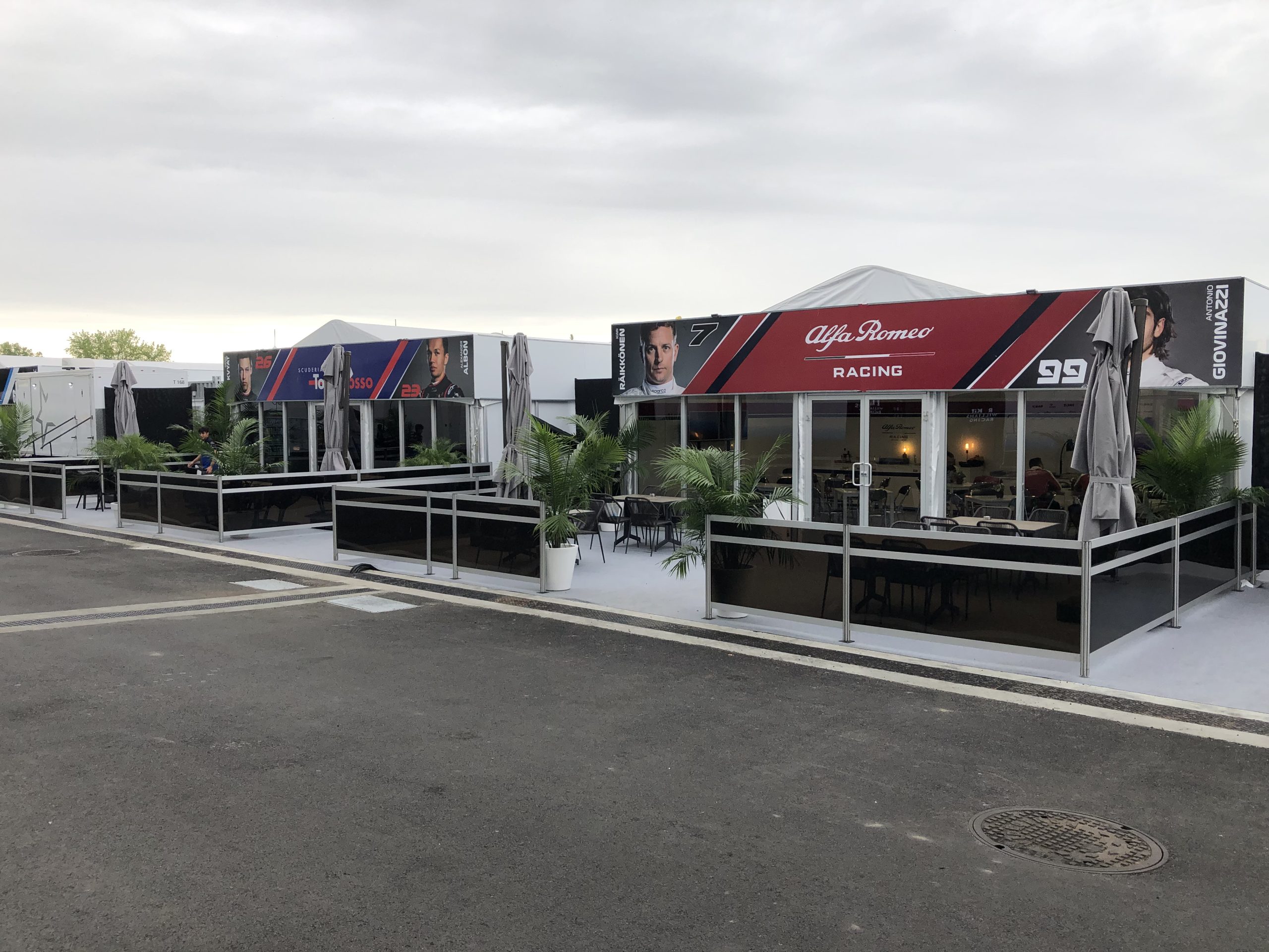F1 Racing Team Hospitality Structures — Grand Prix Paddock Alfa Romeo Racing and Scuderia Toro Rosso F1 hospitality structures with branded graphics, glass walls, and outdoor terraces at Grand Prix paddock