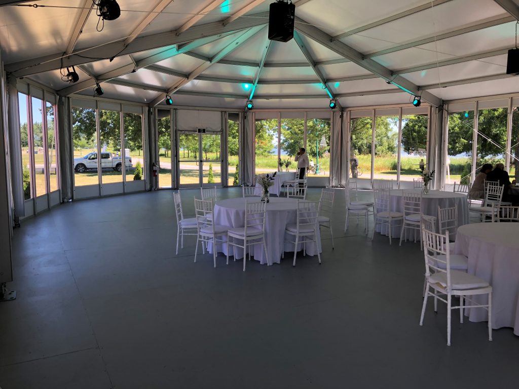 Structure tent interior with floor-to-ceiling glass wall panels, round tables, and chiavari chairs for wedding reception